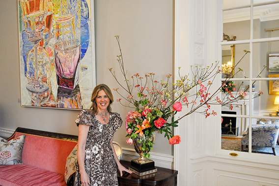 Une femme sourit à côté d'une grande composition florale rose avec des branches qui en sortent. L'art est à l'arrière-plan.