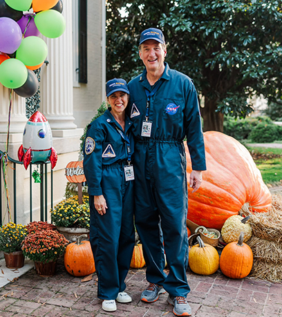 Le gouverneur et la première dame, vêtus de combinaisons d'astronautes de la NASA assorties, posent ensemble en plein air lors d'un événement festif d'automne. Ils se tiennent devant un étalage présentant une grosse citrouille, des bottes de foin et des ballons colorés, avec une petite fusée jouet à proximité. Le cadre comprend une allée en briques et un bâtiment classique à colonnes blanches, entouré de décorations saisonnières.