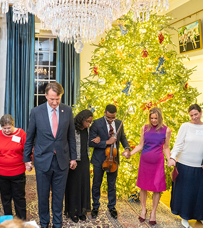 Un groupe de six personnes est debout en demi-cercle, se tenant par la main devant un sapin de Noël illuminé et orné de boules bleues et rouges. Le cadre semble être une grande pièce élégamment décorée avec un lustre et un tapis à motifs recouvrant le sol. Sur le côté gauche, de longs rideaux bleus froncés encadrent une fenêtre. Les individus sont vêtus d'une variété d'atours, allant de l'uniforme à l'uniforme, en passant par le costume.