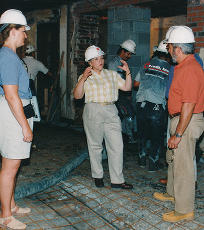 La première dame, Roxane Gilmore, porte un casque de protection dans une structure inachevée et discute avec deux autres personnes portant des casques de protection. Mme Gilmore porte une chemise boutonnée à manches courtes et un pantalon kaki. Cette photo a été prise entre 1998-2000.