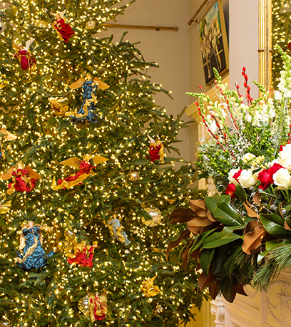 Contexte : Un grand arbre est décoré pour Noël avec des lumières blanches, des ornements en verre transparent et des anges portant des robes rouges, bleues et dorées. Premier plan : Sur le côté droit de l'image, un manteau est décoré de roses rouges et blanches et de verdure avec des lumières blanches chaudes.