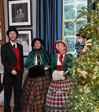Trois artistes en tenue d'époque se tiennent à côté d'un arbre de Noël illuminé. Certains ont la bouche ouverte, ce qui laisse penser qu'ils chantent.