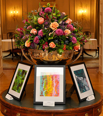 Trois tableaux dans des cadres noirs sont posés sur une table ronde avec, derrière eux, une composition de fleurs roses.