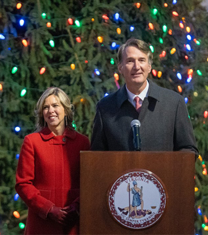Le gouverneur Youngkin et la première dame Suzanne Youngkin se tiennent derrière un podium devant un grand arbre de Noël aux lumières multicolores.