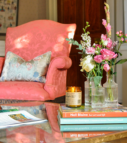 Une table basse avec trois vases à fleurs et une bougie posée sur une pile de livres. Un fauteuil corail avec un coussin décoratif se trouve à l'arrière-plan.