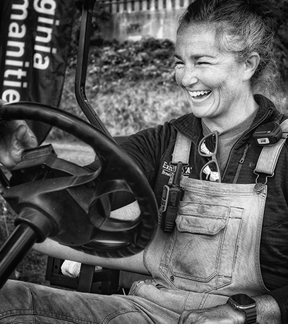 Une personne sourit, assise dans un véhicule, vêtue d'une salopette et munie d'une radio. Photo en noir et blanc.