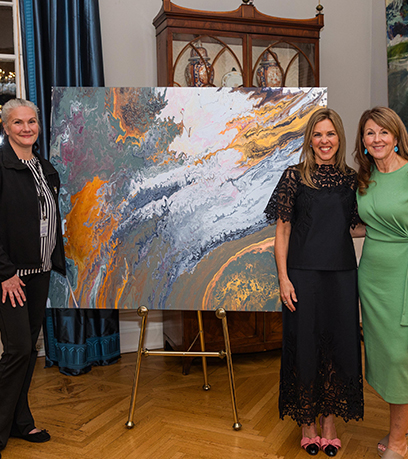 Trois femmes debout à côté d'une peinture abstraite aux motifs tourbillonnants dans les tons orange, blanc, gris et vert.