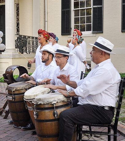 La Tradicion Dancy Company se produit à l'extérieur de l'Executive Mansion. Trois hommes assis jouent du tambour au premier plan et un groupe de femmes chante derrière eux.
