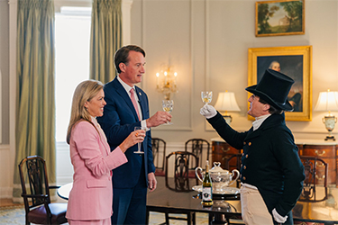 Une femme en costume rose, un homme en costume bleu avec une cravate rose et un homme vêtu d'habits du XVIIIe siècle lèvent leur verre pour porter un toast.