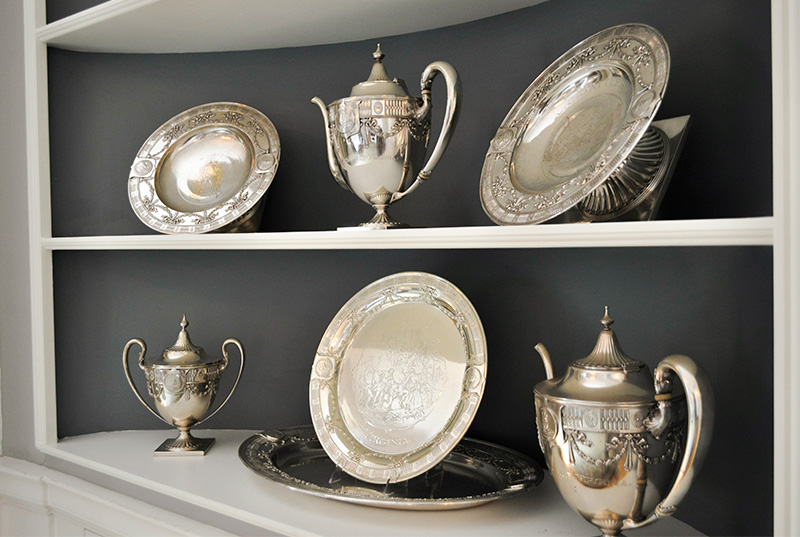 Six pièces d'argenterie (trois théières et trois assiettes) reposent sur une étagère.